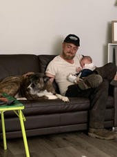 A man is sitting on a couch holding a baby and a dog.