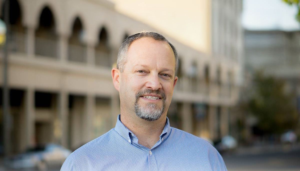 A man with a beard is smiling for the camera in front of a building.