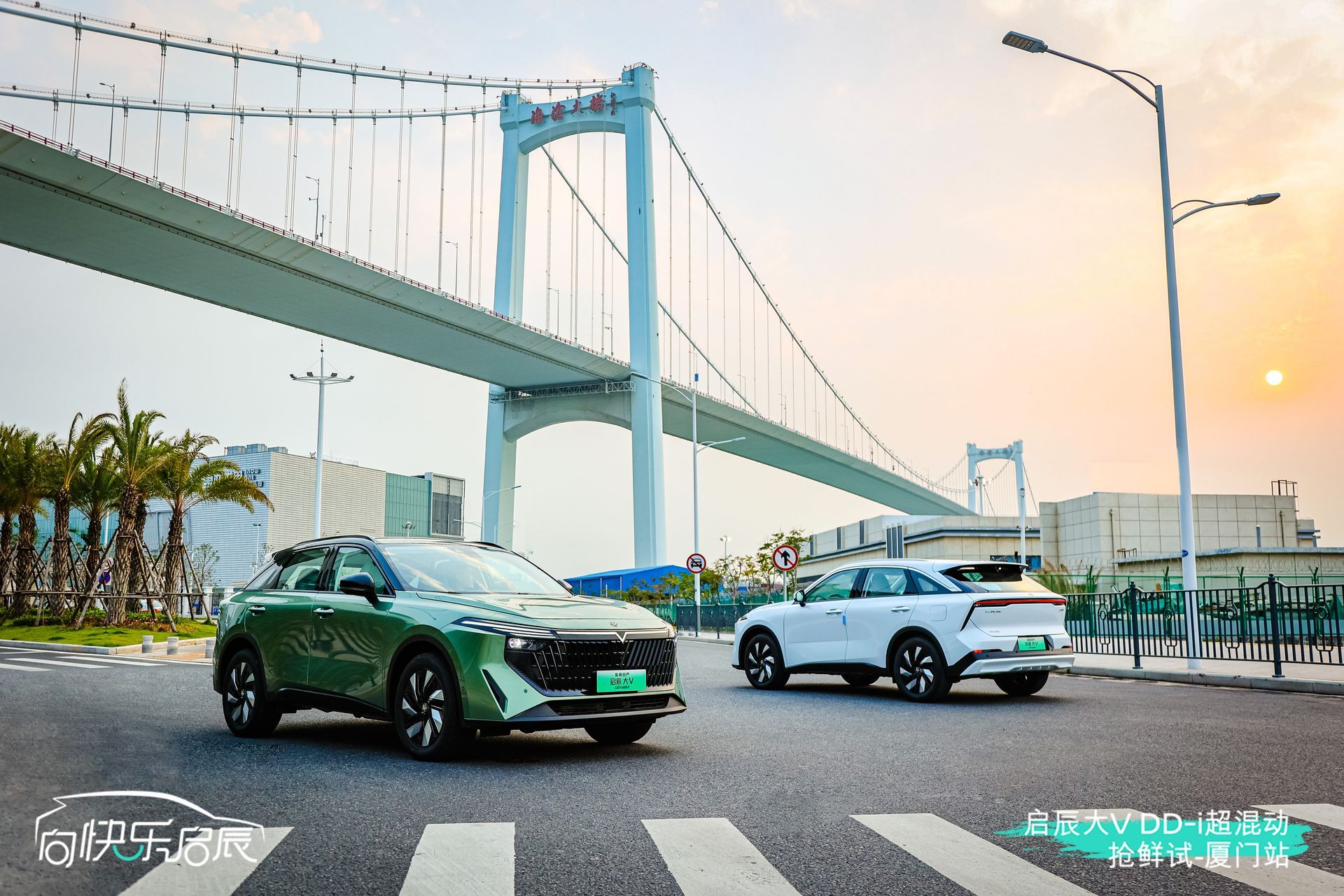 Two electric SUVs on a road under a suspension bridge; one green, one white. Sunset in the background.