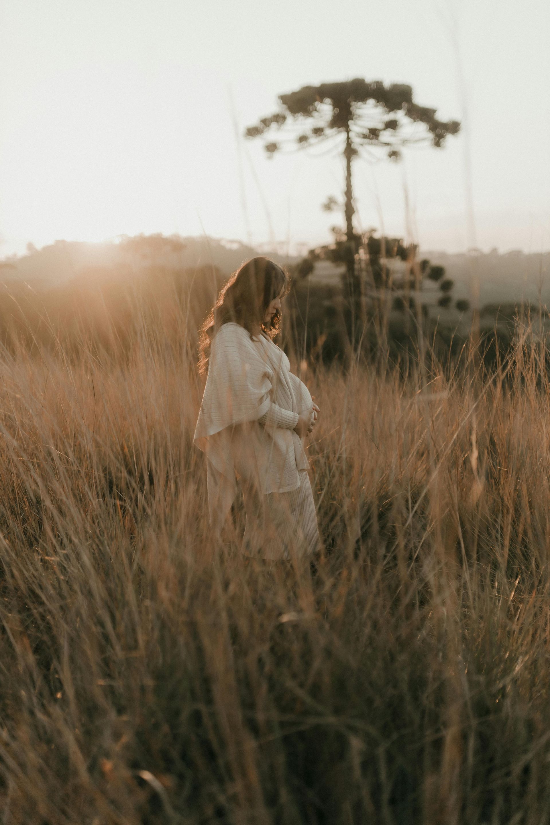 Woman in white dress stands in tall, dry grass field, facing away, with tree in background at sunset.