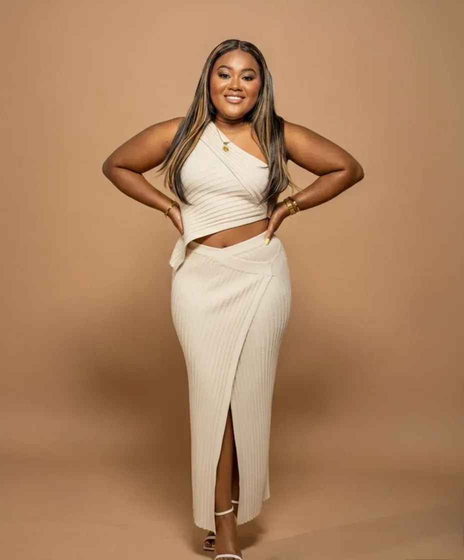 Woman wearing a cream-colored one-shoulder top and skirt, posing with hands on hips, neutral background.