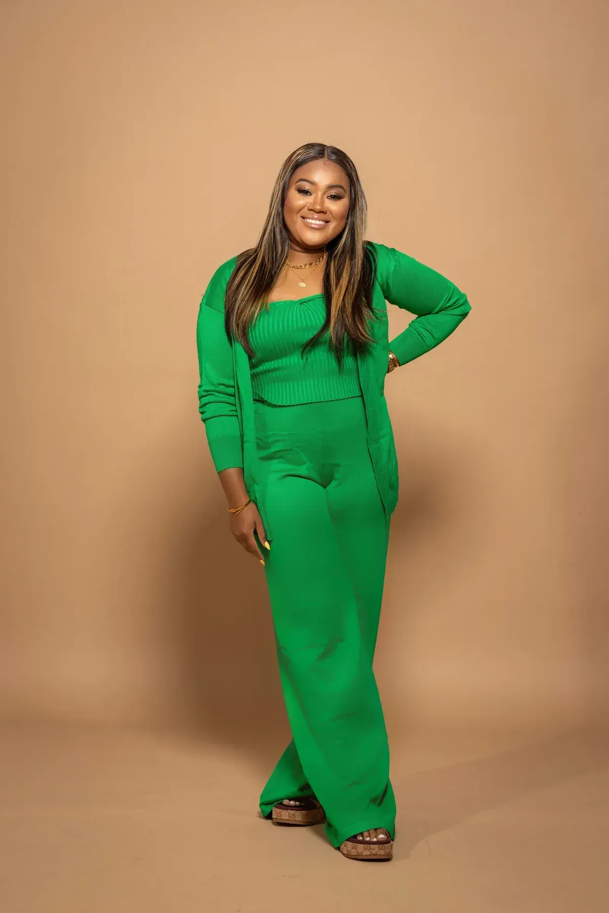 Woman in a green knit outfit, smiling, hand on hip, standing against a tan backdrop.