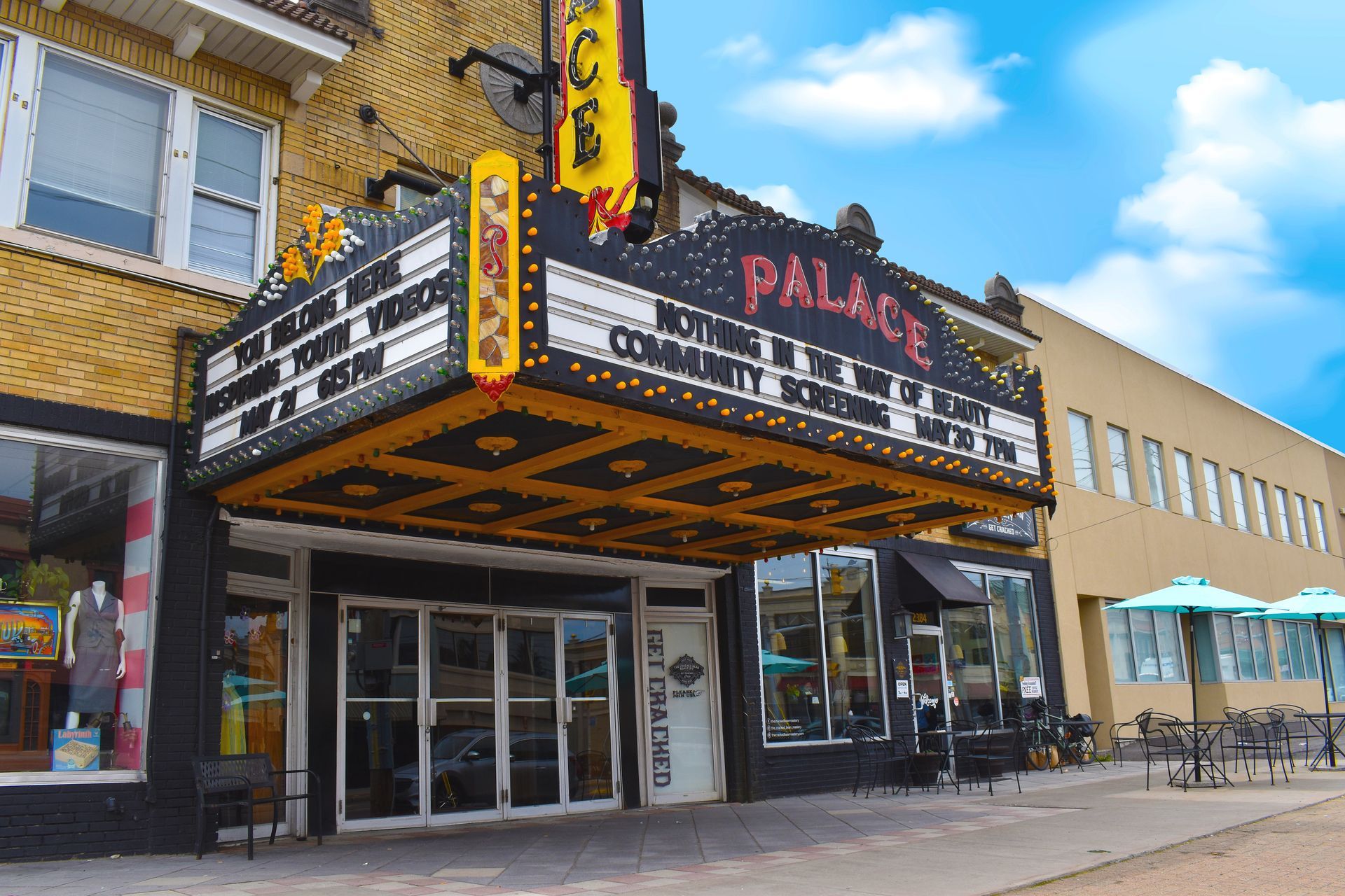 The Palace Theatre: marquee with movie titles, black facade, awning, brick and tan buildings, blue sky.