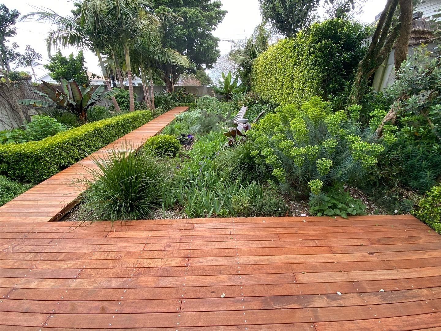 A wooden walkway in a garden surrounded by trees and bushes.