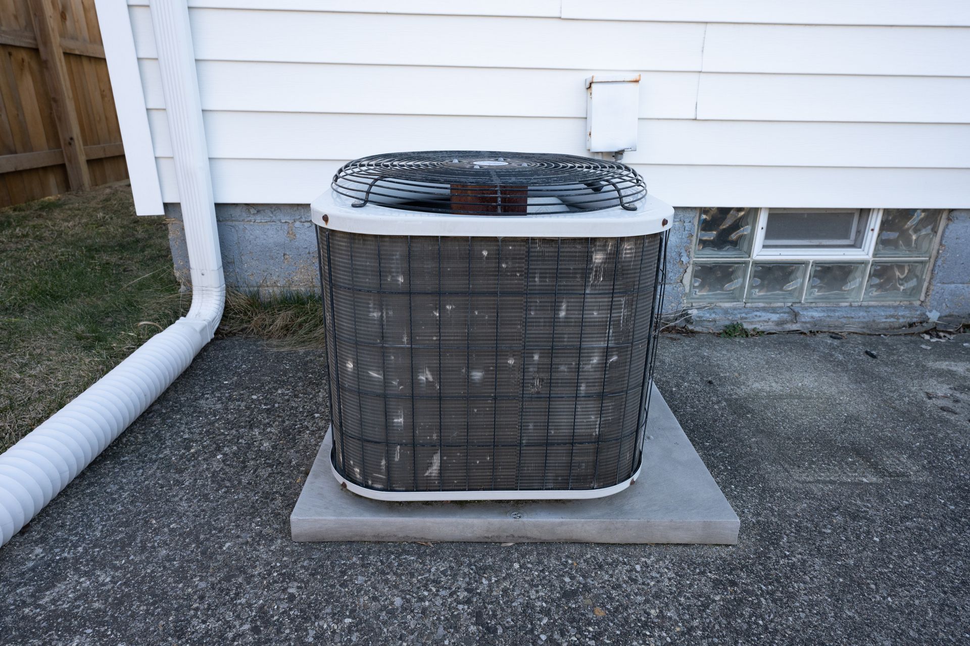 An outdoor residential air conditioning condenser unit sitting on a concrete pad against a white-sided house.