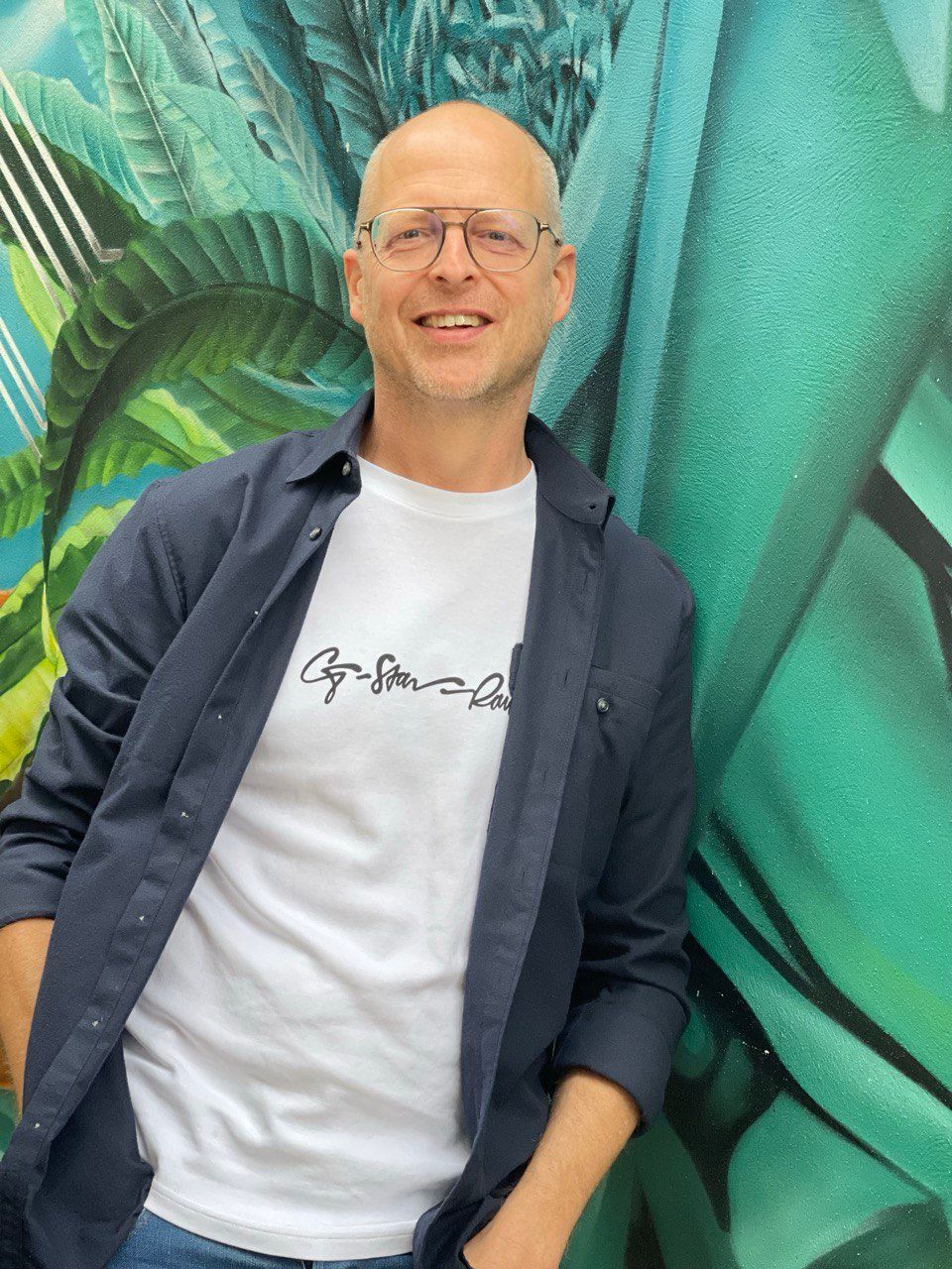 Man with glasses smiles, leaning against a teal mural. He wears a white shirt, blue open button-up, and jeans.