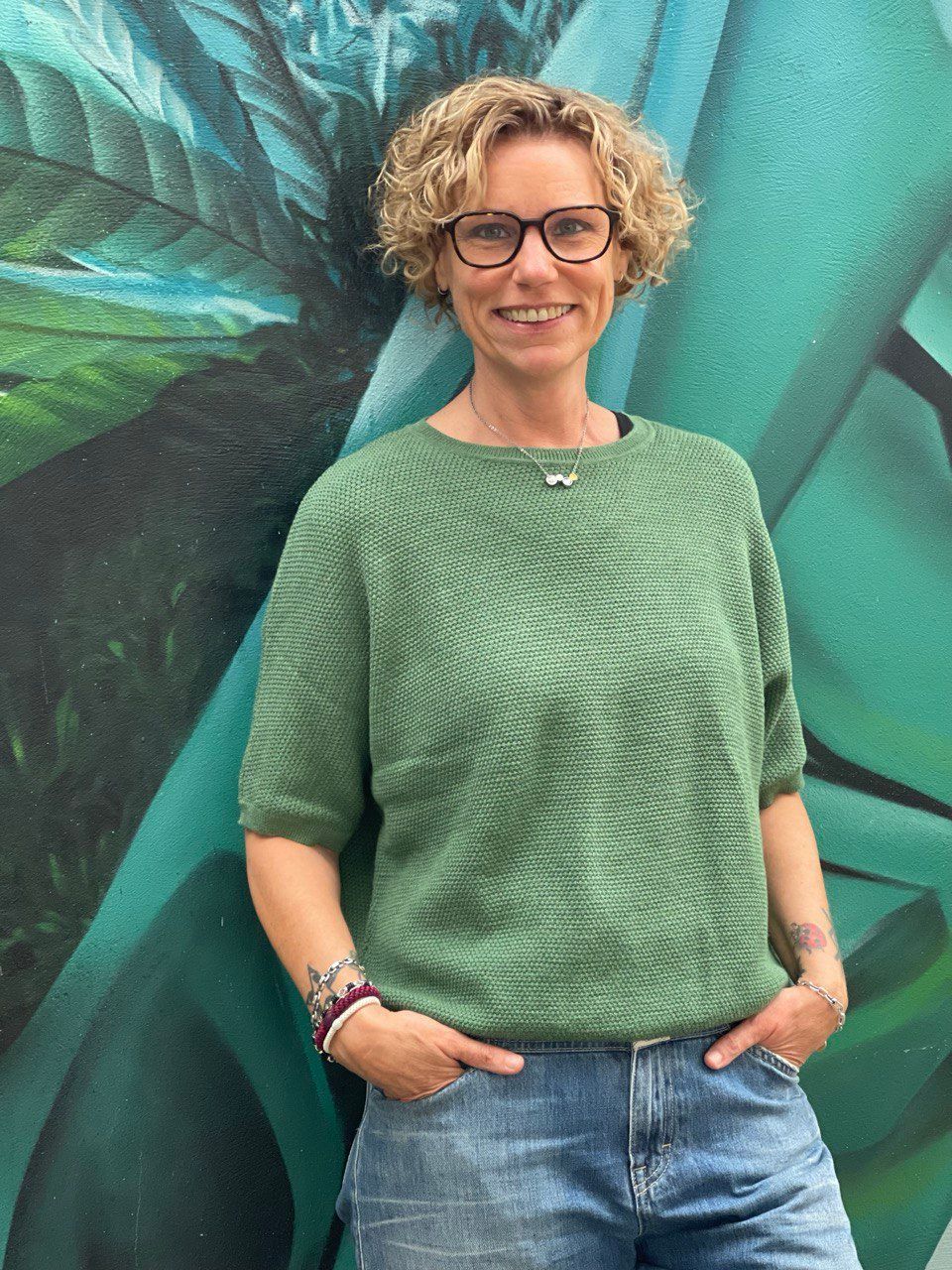 Woman with curly hair in green shirt and jeans, hands in pockets, smiling, against a green mural.