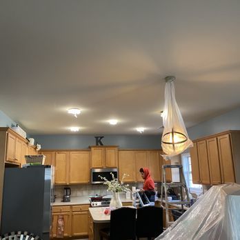 Person on ladder working on a kitchen light fixture; light wood cabinets and grey appliances.
