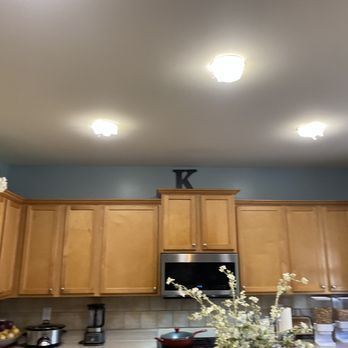 Kitchen with light wood cabinets, stainless steel appliances, blue wall, and overhead lights. A "K" hangs above the microwave.