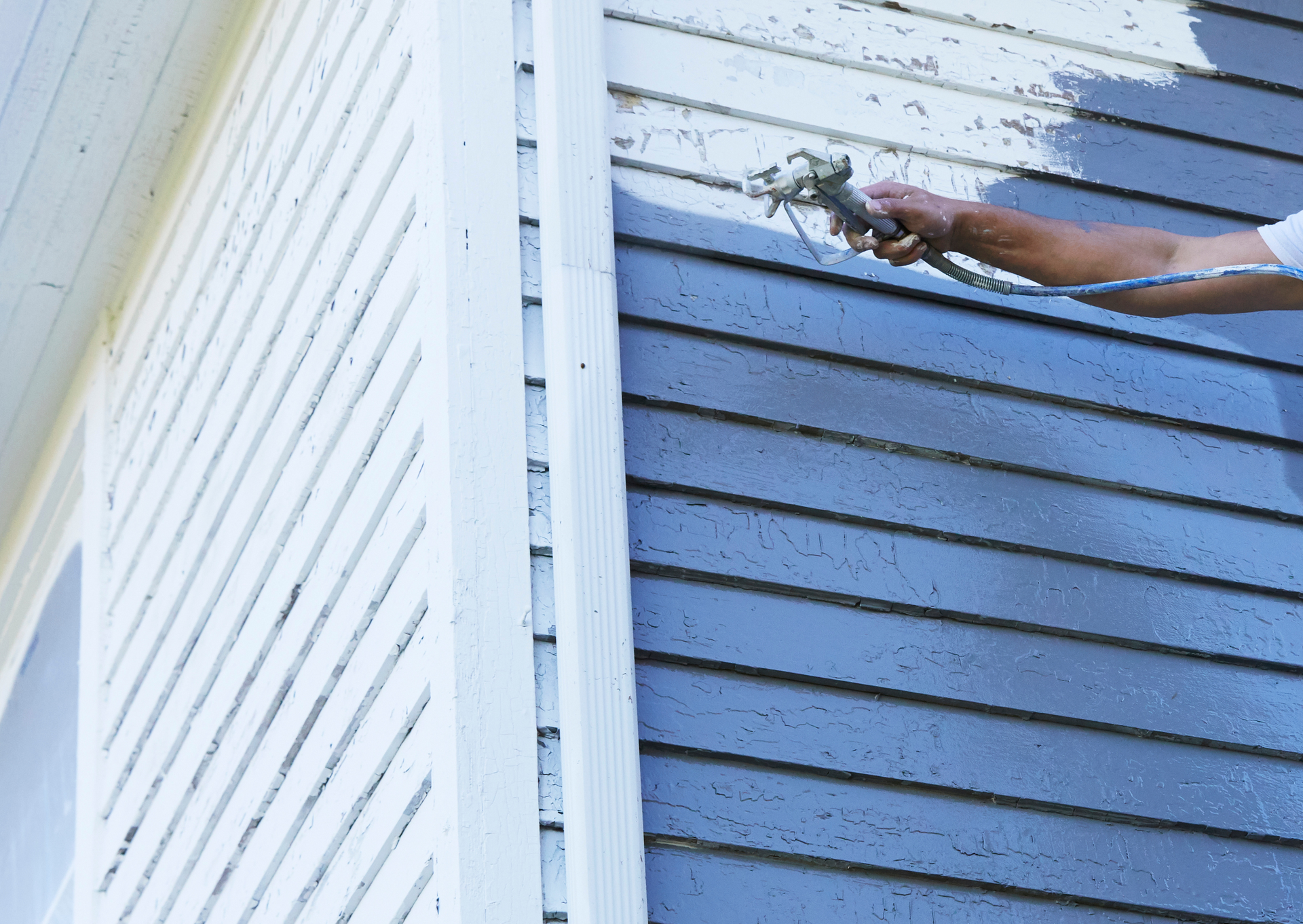 Person spraying paint onto a blue-gray house siding.