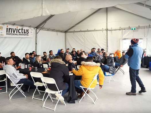 A group of people seated at tables inside a tent, listening to a presentation. A man stands at the front speaking.