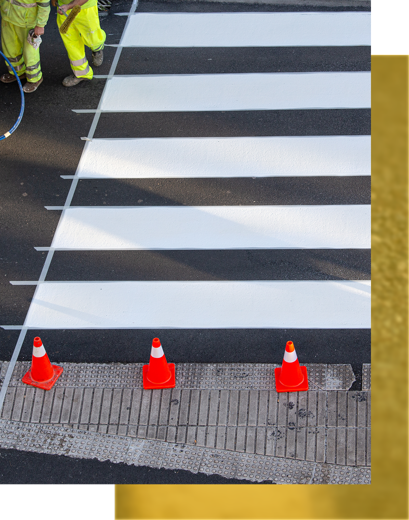 Workers painting a crosswalk, using a sprayer. White stripes on asphalt. Orange cones set up.