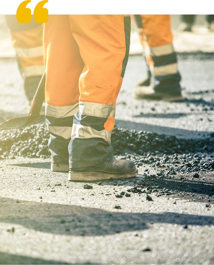 Snowplow clears snow from a road in front of a yellow building and trees.