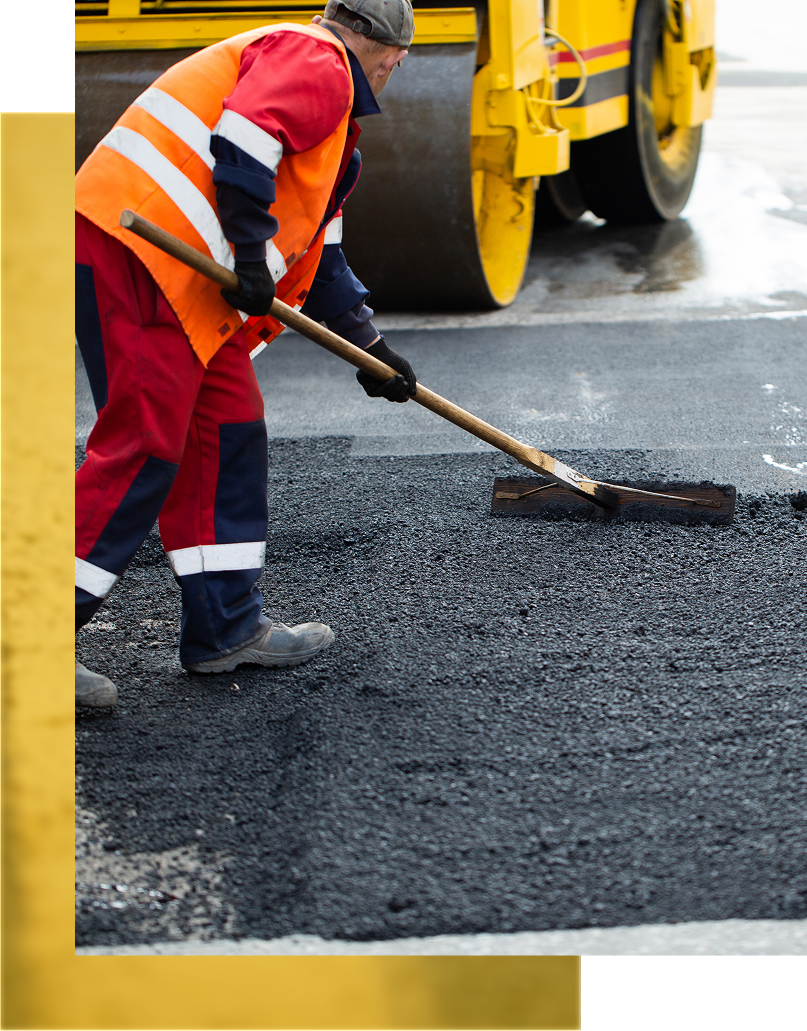 Road worker smoothing asphalt with a broom, roller in background.