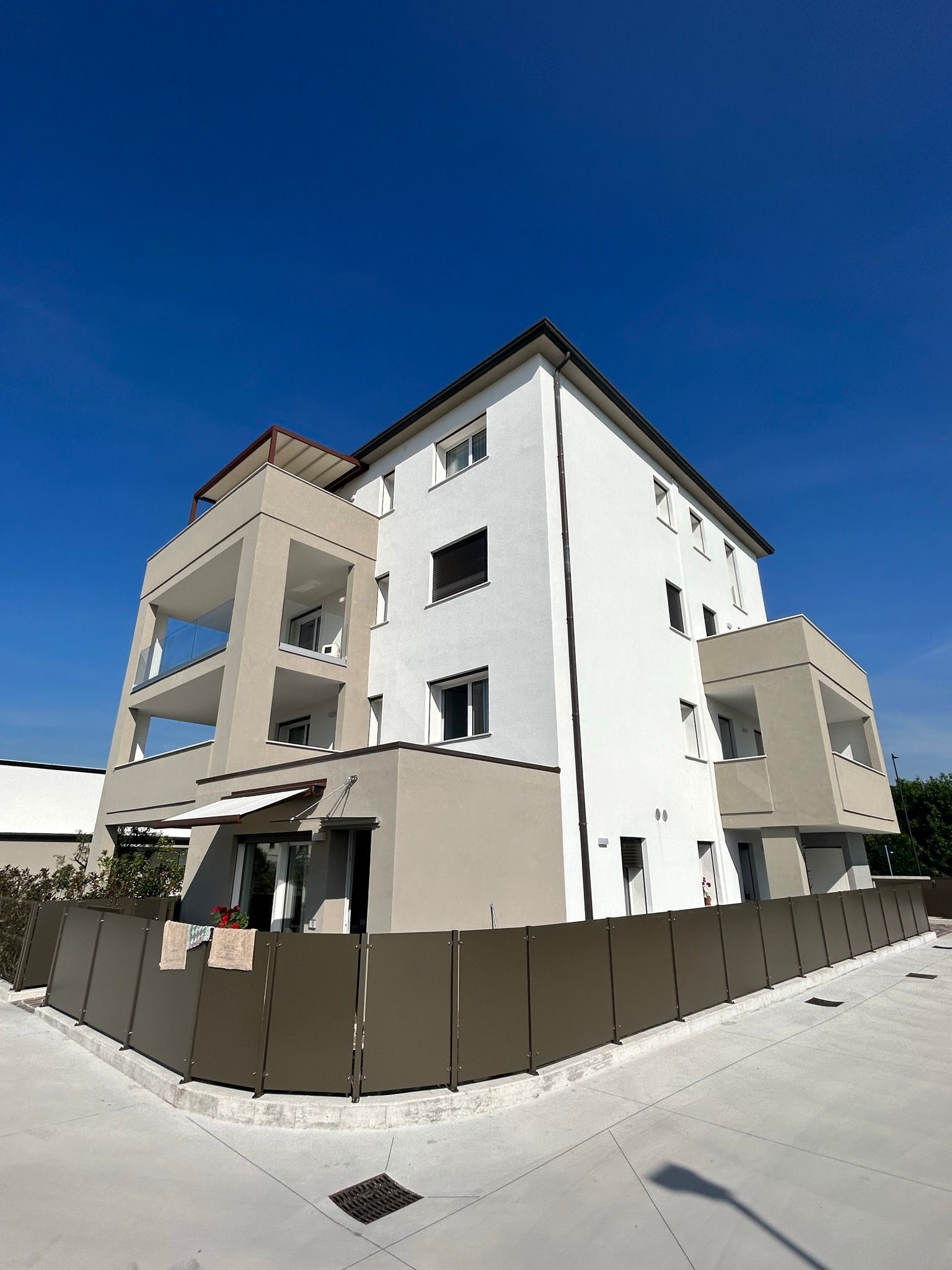 Moderno condominio con facciate bianche e beige, situato dietro un lotto d'angolo recintato sotto un cielo azzurro limpido.