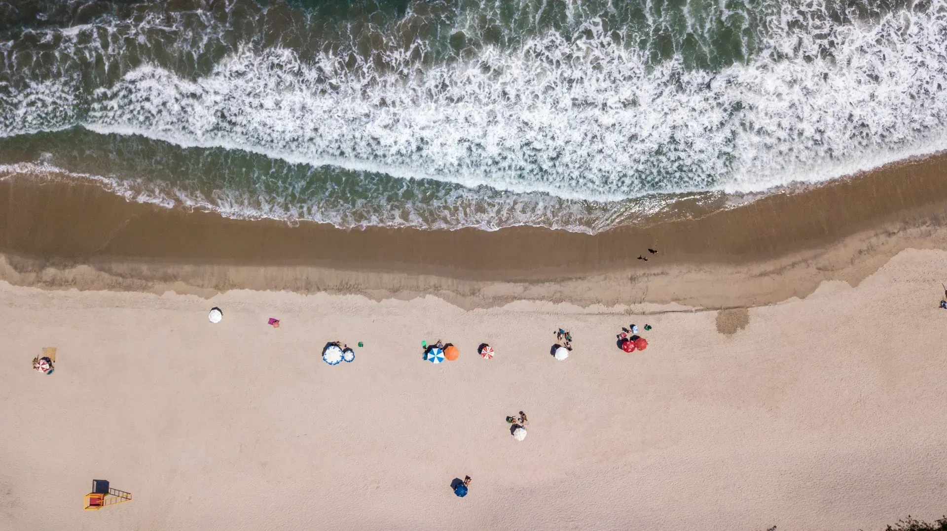 Uma vista aérea de uma praia com pessoas sentadas na areia e ondas quebrando na costa.