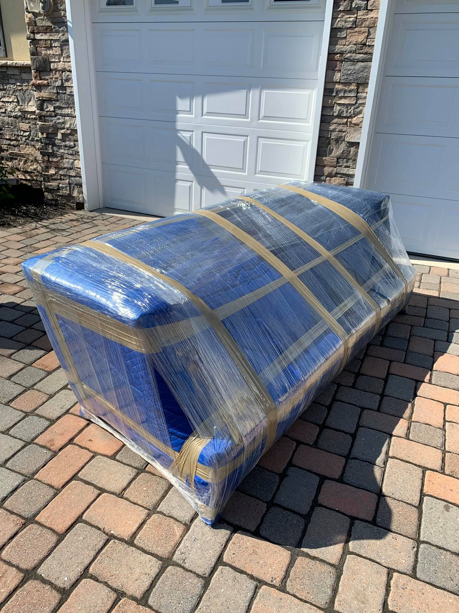 A blue couch wrapped in plastic is sitting on a brick driveway in front of a garage.