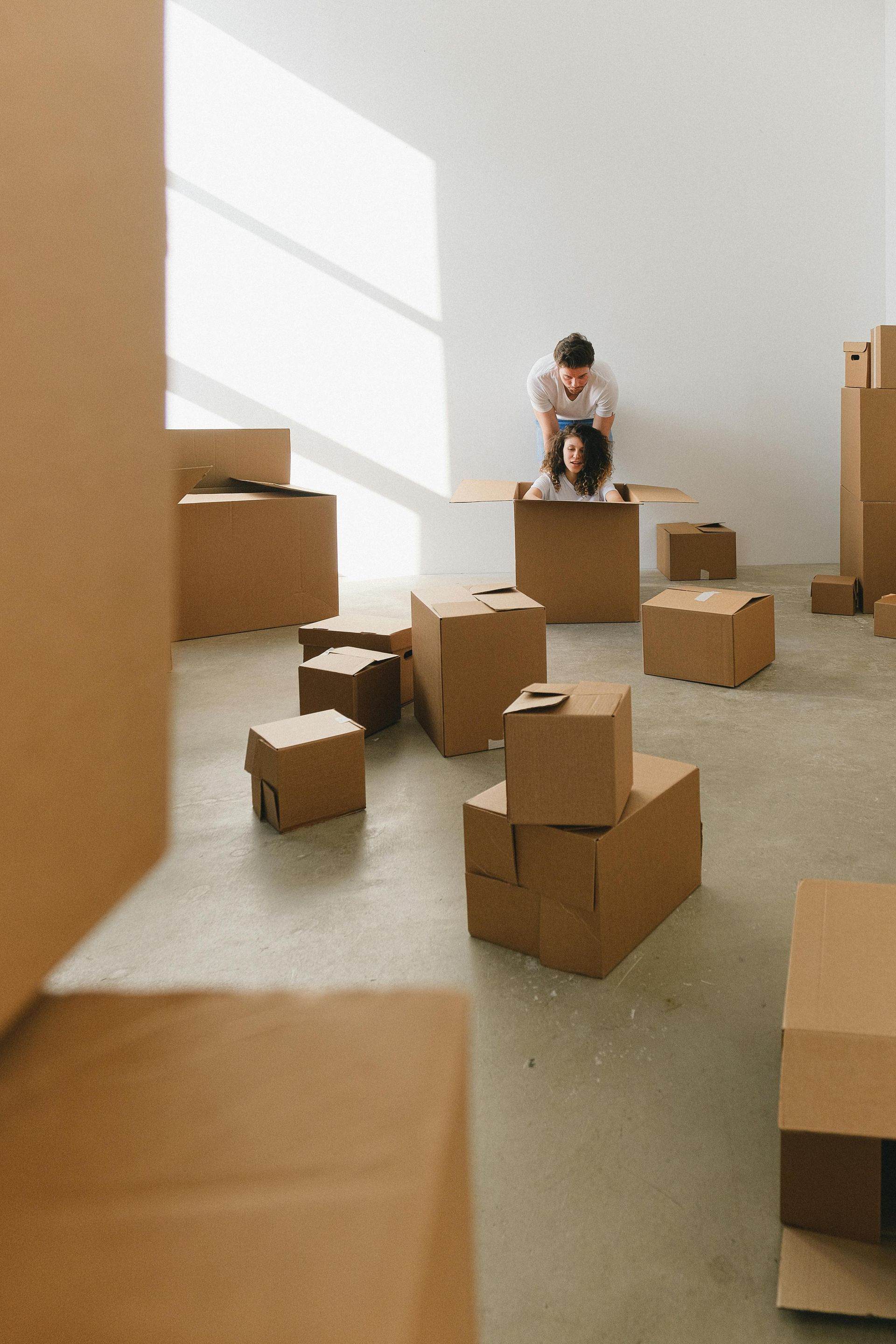 Couple playing with cardboard boxes in a room, one inside a box, the other pushing.
