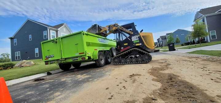 A green dumpster is being pulled by a bulldozer on a dirt road.