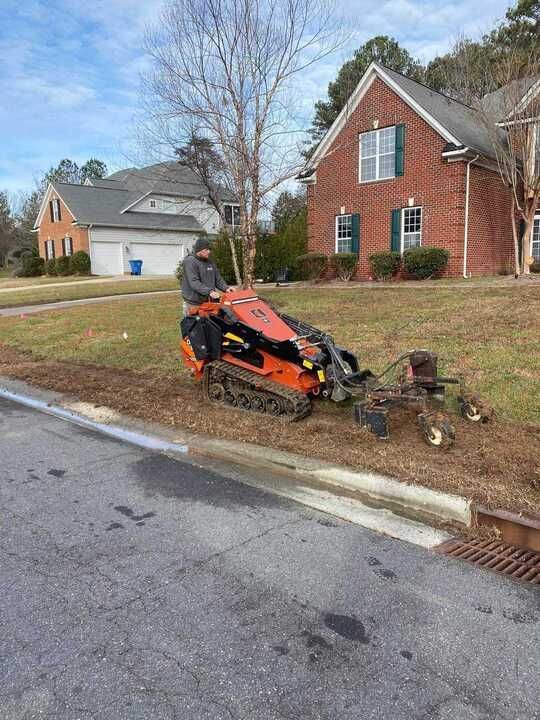 A man is driving a tractor down a street next to a brick house.