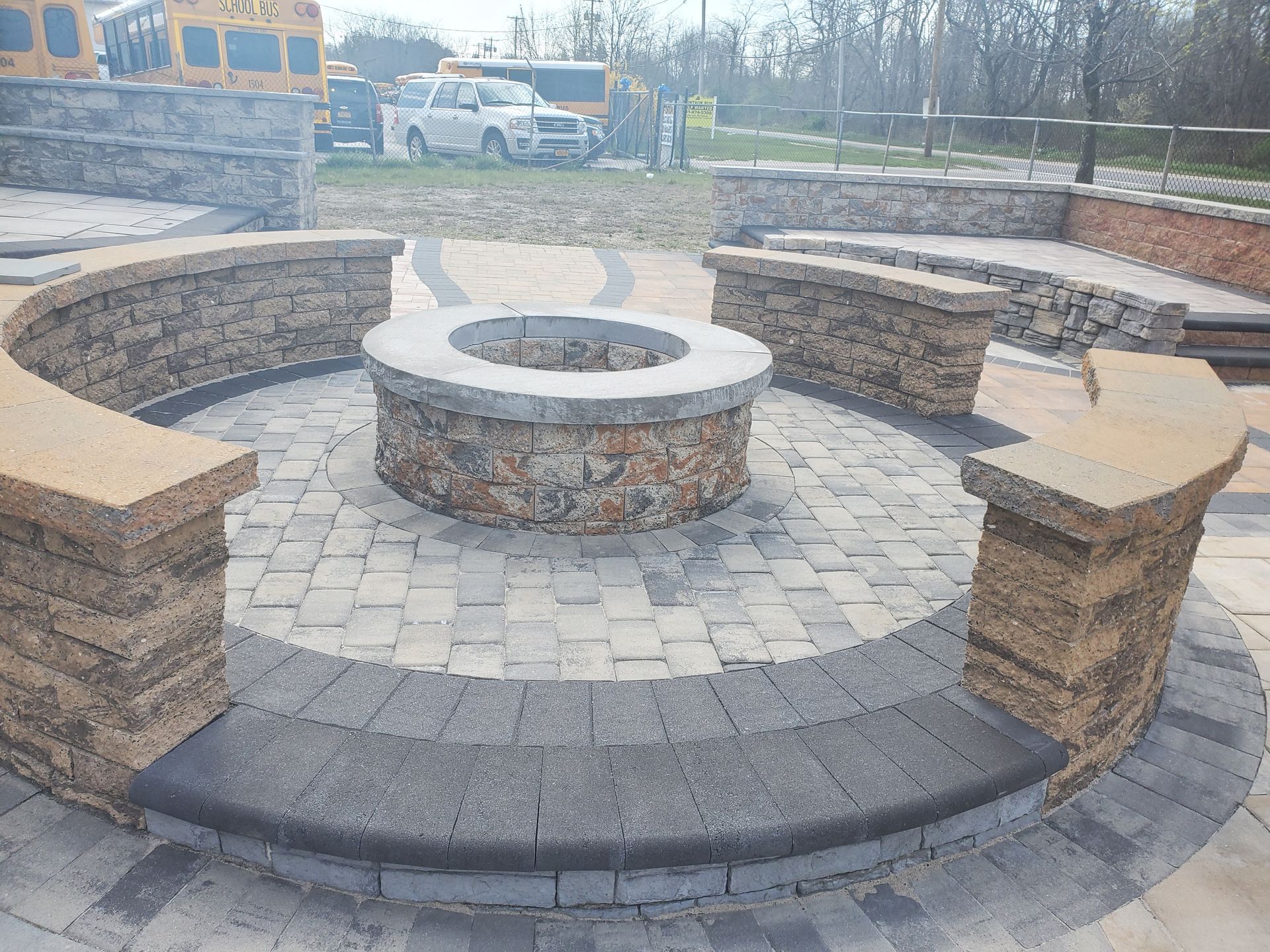 A fire pit in the middle of a brick patio with a school bus in the background.