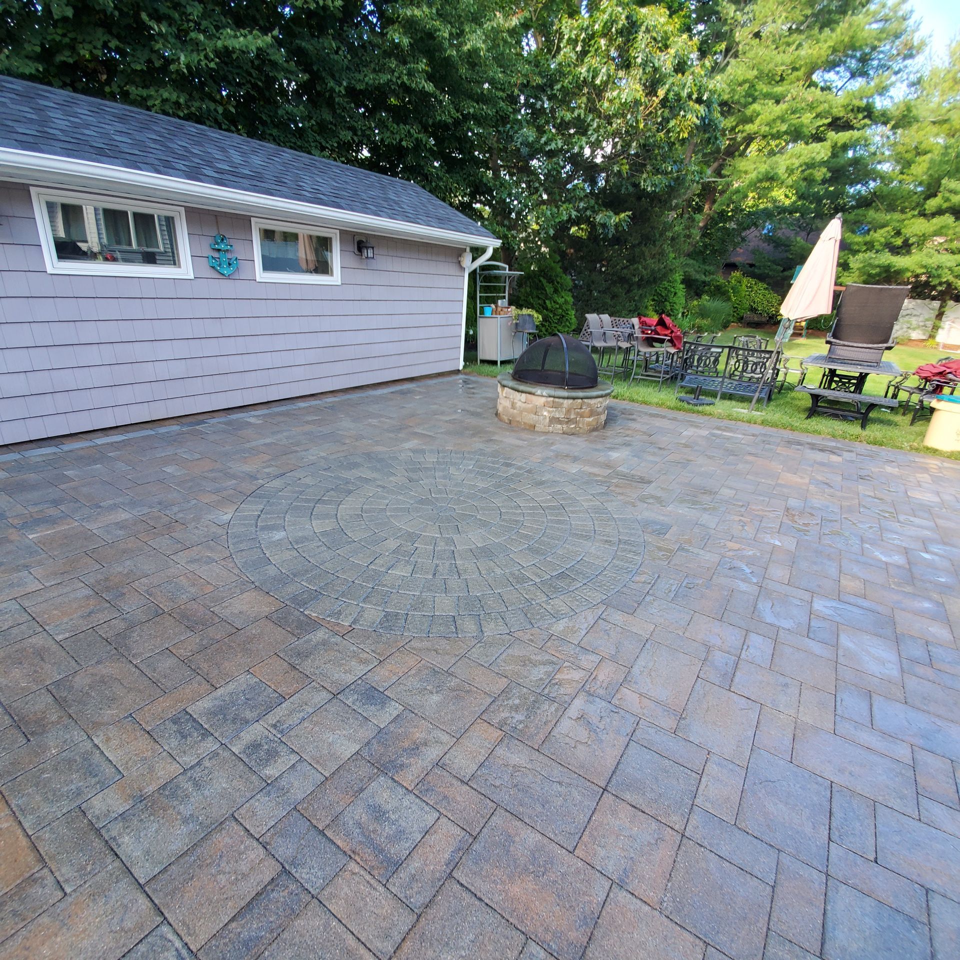 Patio with brick pavers, fire pit, and outdoor furniture next to a gray building and trees.