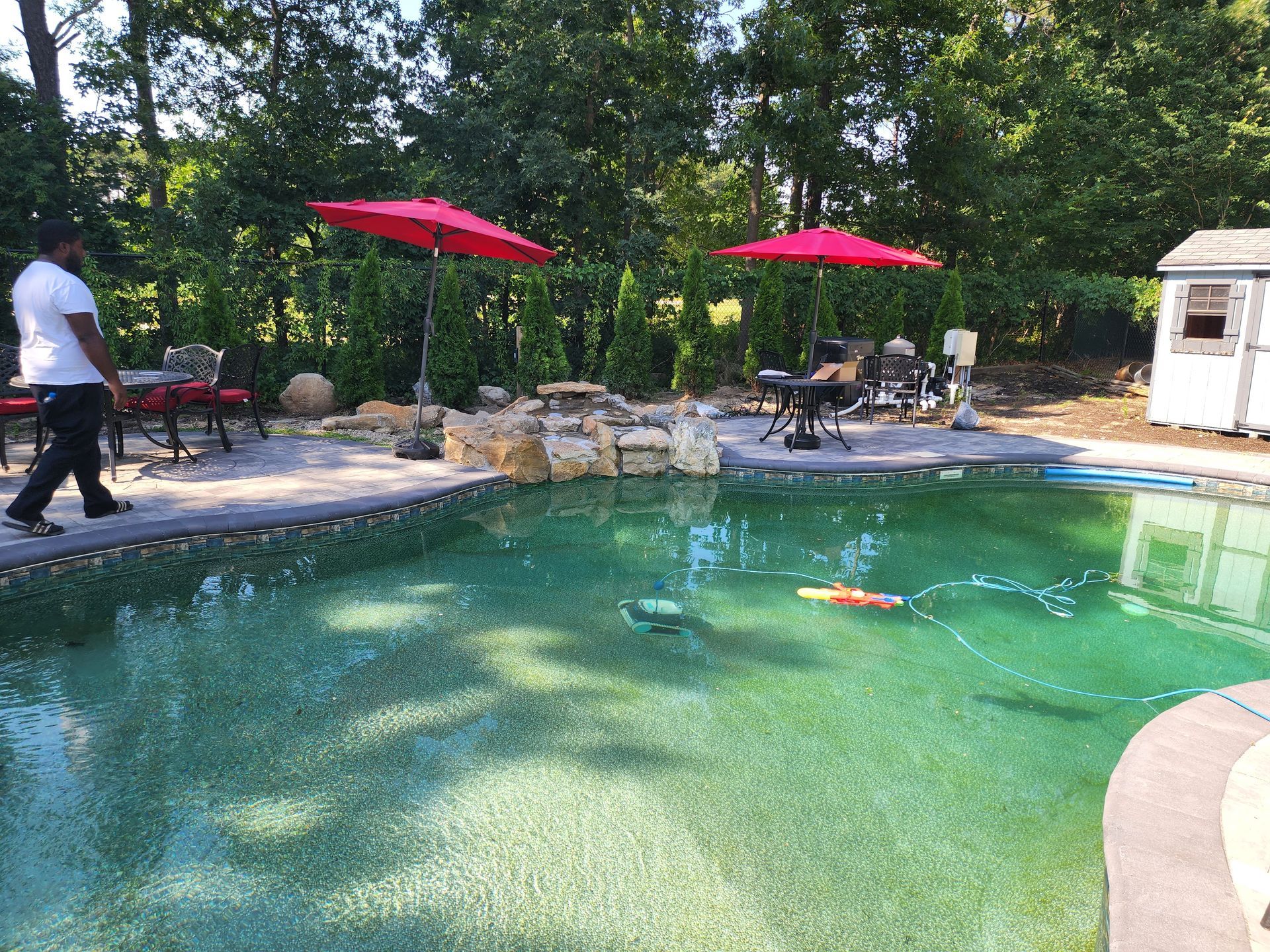 A man is standing next to a swimming pool with red umbrellas.