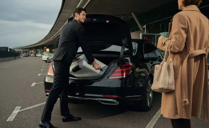 Man in a suit loading luggage into a black car trunk at an airport. A woman in a coat stands nearby.