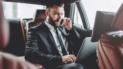 A businessman in a suit is in the back of a car, using a laptop and talking on the phone.