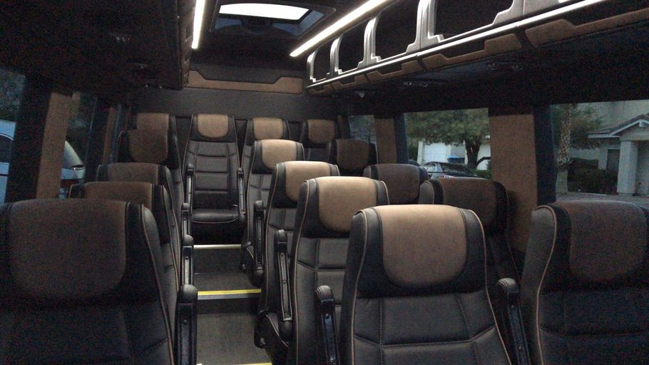 Interior of a passenger van with rows of brown and black seats, overhead storage, and interior lighting.