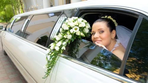 Bride in a white limo with a bouquet, smiling, looking out window.