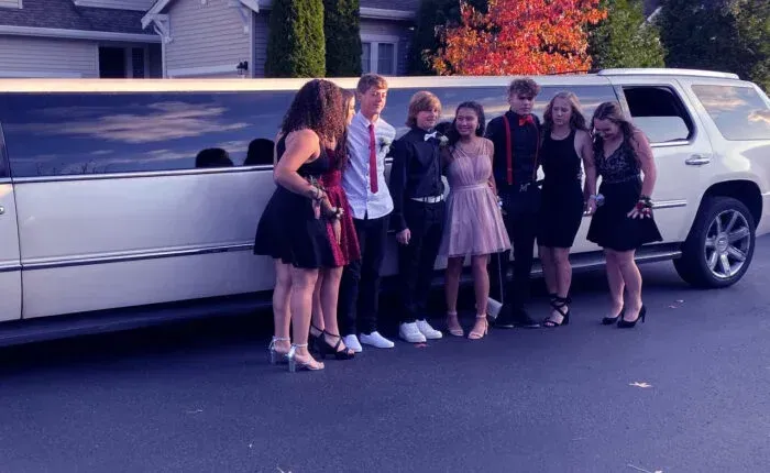 Group of teens in formal wear pose by a white limousine outside; autumn colors in the background.