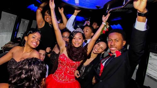 Teens in formal attire dance inside a party bus; joyful expressions, arms raised, a red dress, and a tiara.
