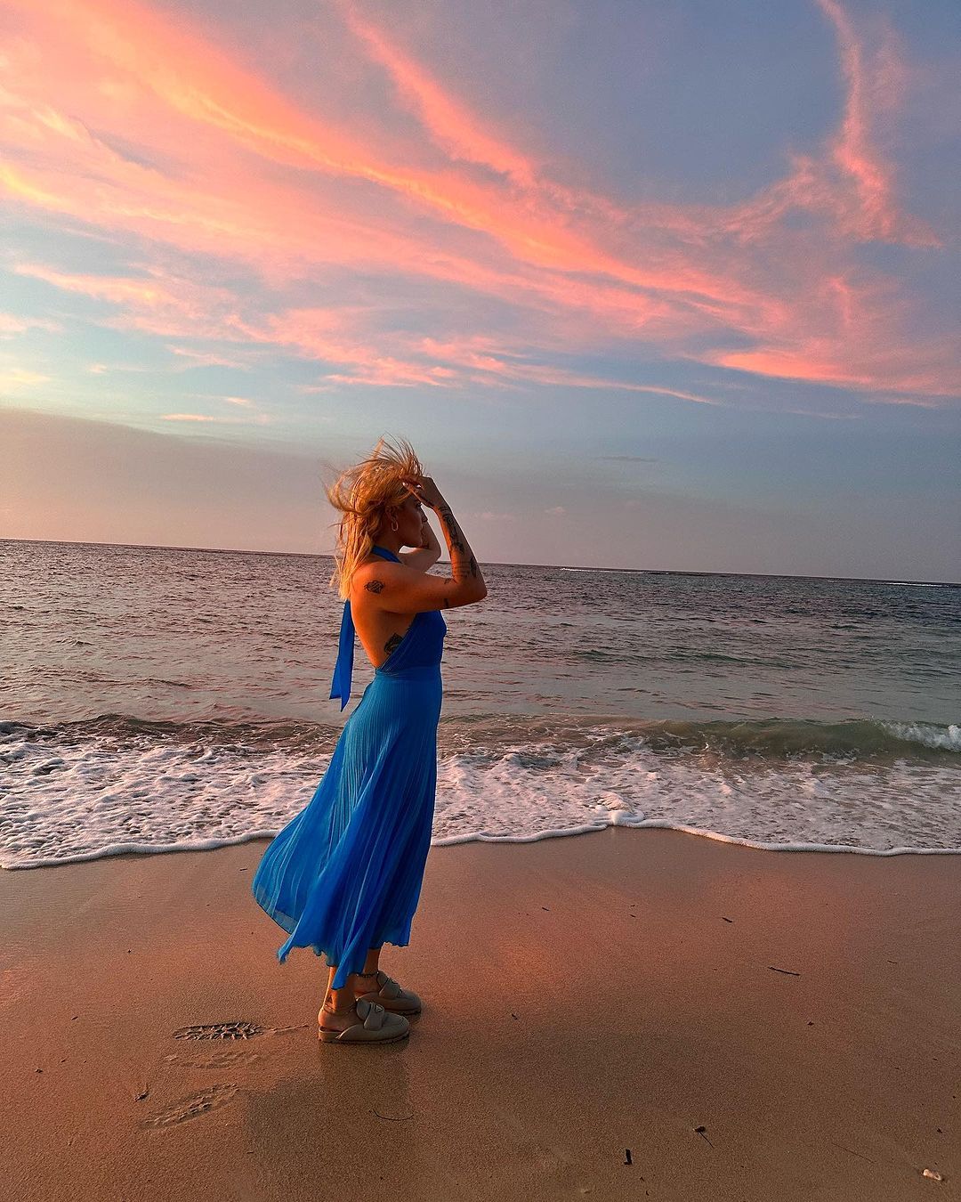 A woman in a blue dress is standing on the beach at sunset.