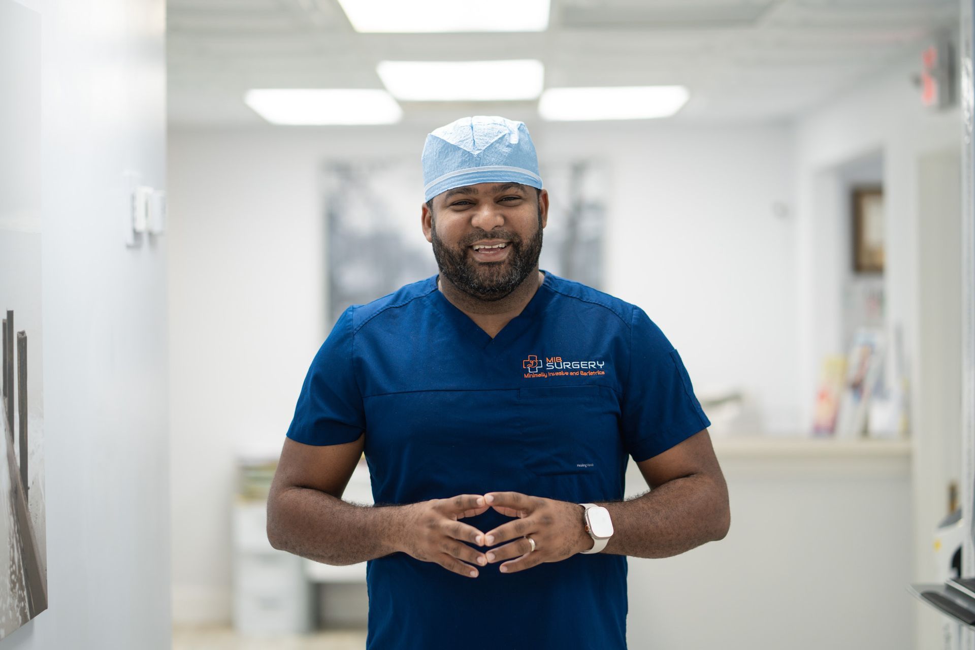 A man in a blue scrub top and surgical cap is standing in a hallway.