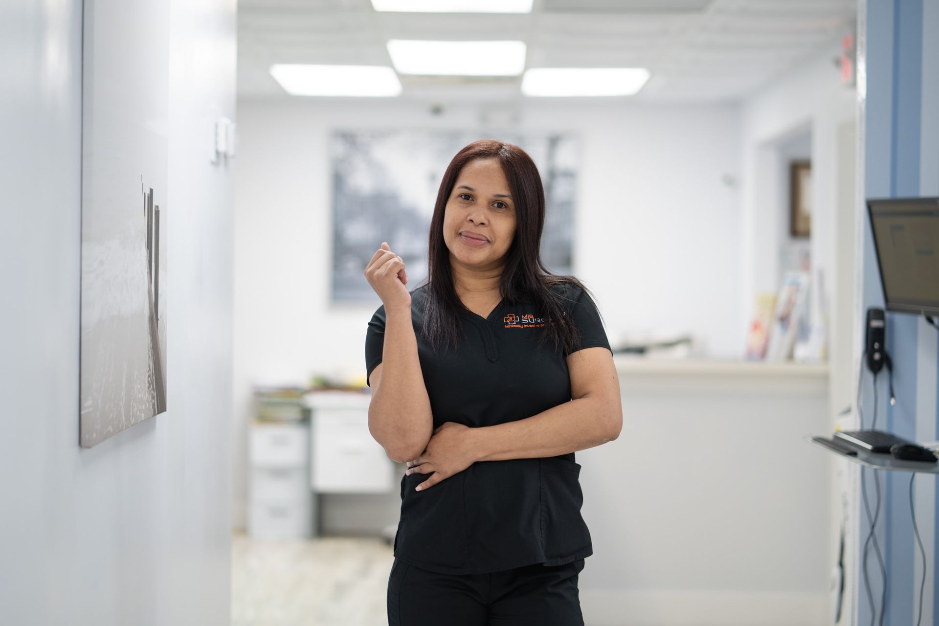 A woman in a black scrub top is standing in a hallway with her arms crossed.