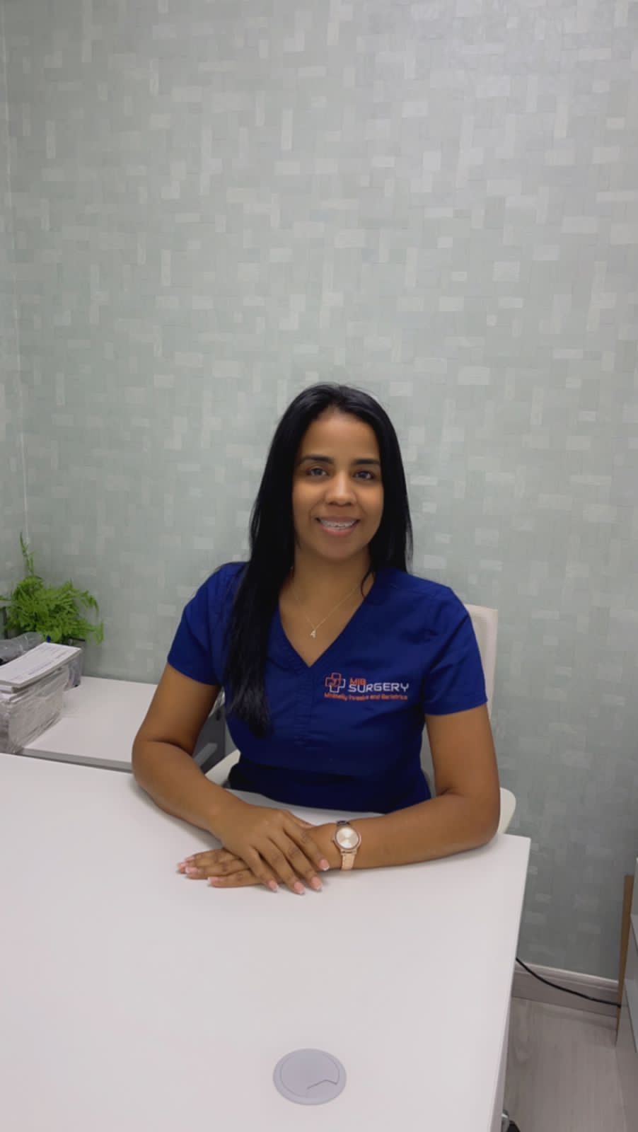 A woman in a blue scrub top is sitting at a desk.