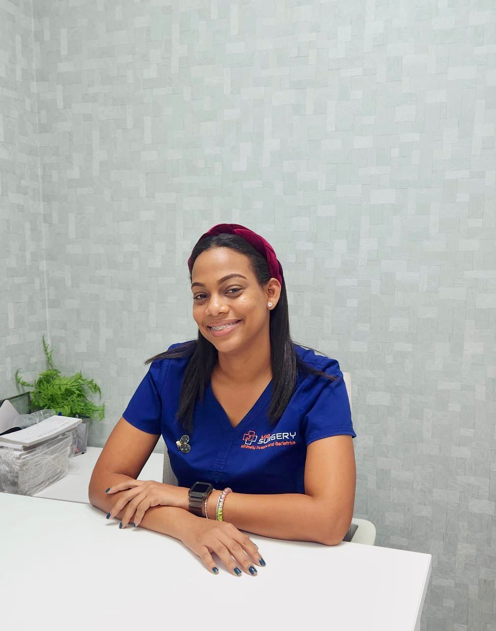 A woman in a blue scrub top is sitting at a desk.