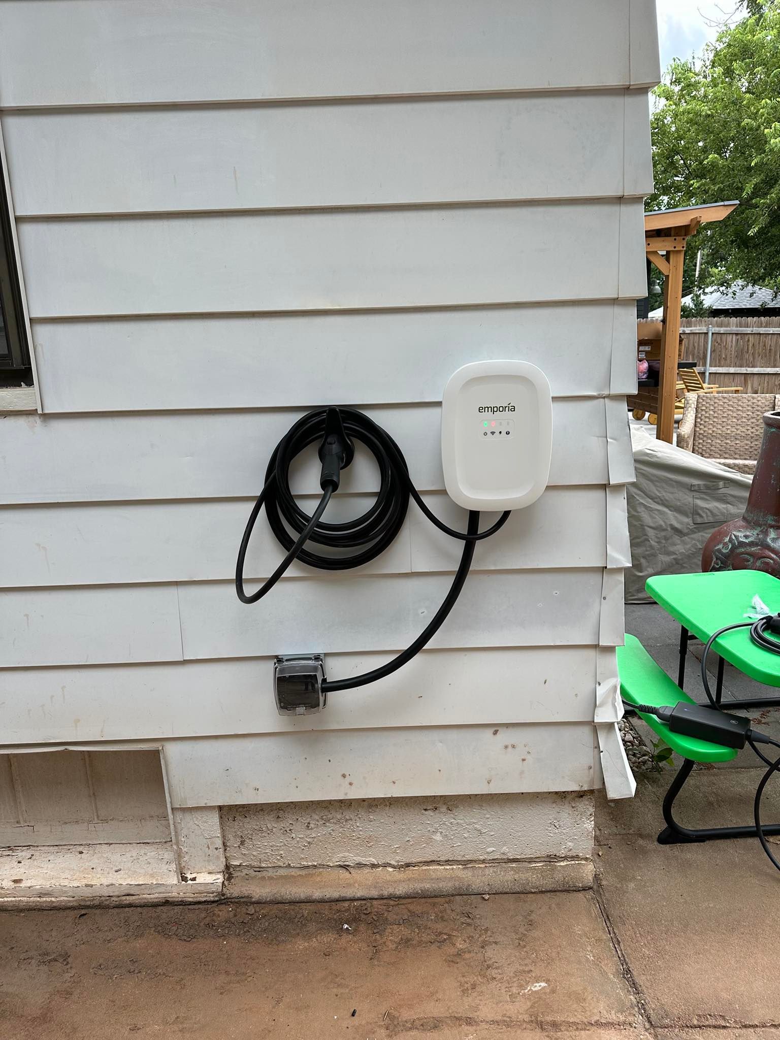 Electric vehicle charging station mounted on the exterior wall of a white house with a coiled black charging cable.