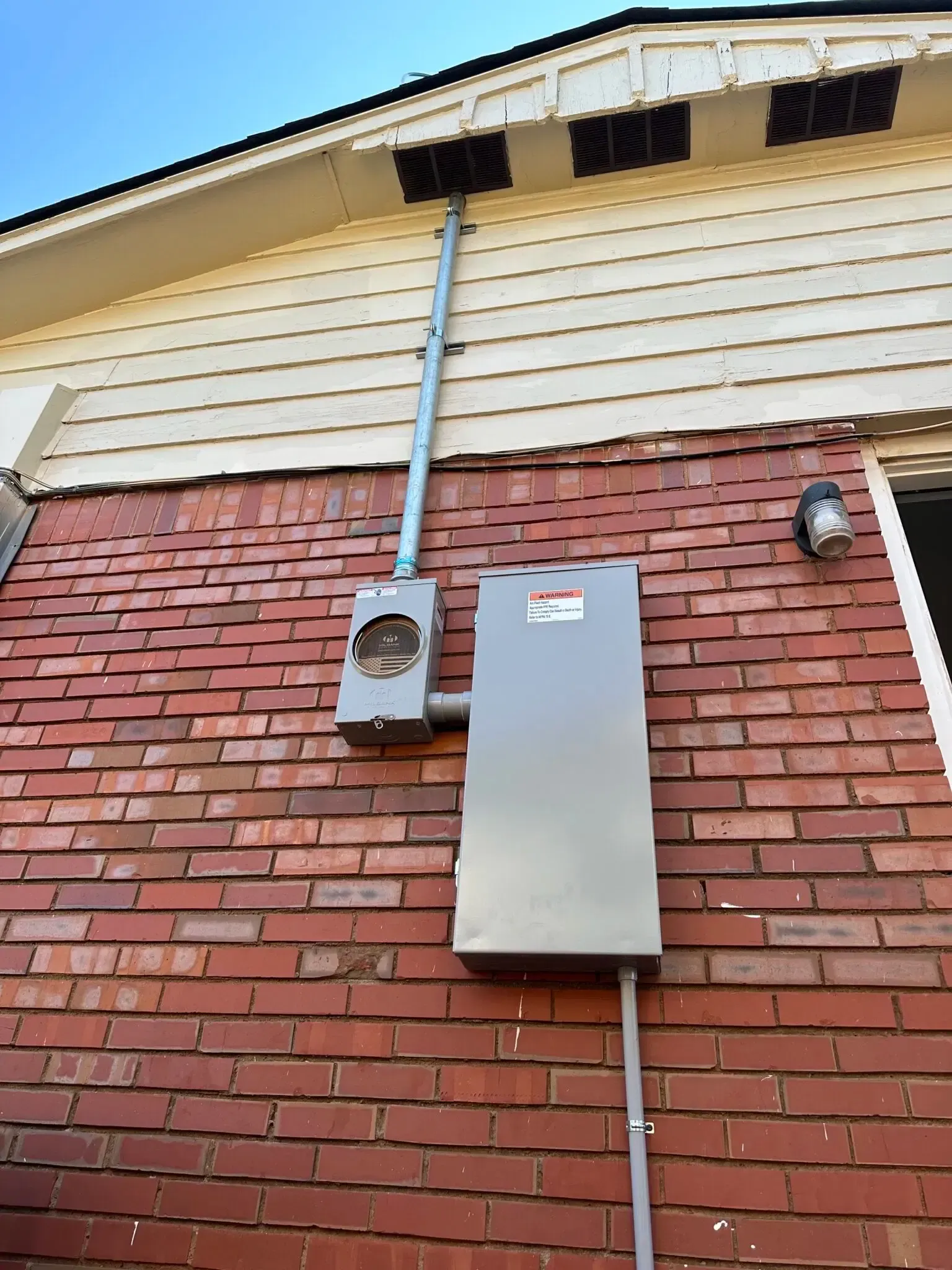 An outdoor electrical meter and breaker box mounted on a brick wall, with a conduit pipe running up to the roof eaves.