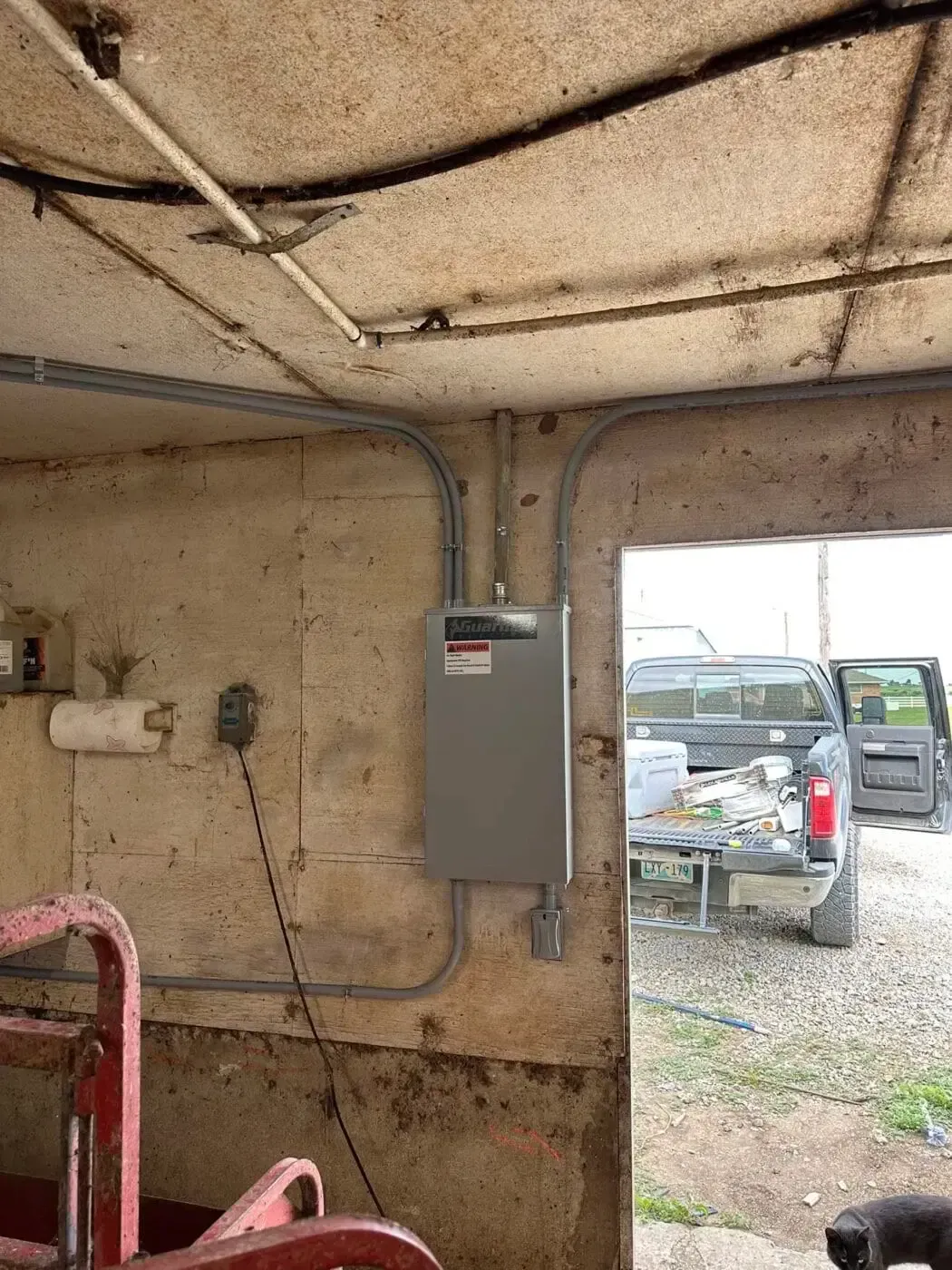A gray electrical subpanel mounted on a cinder block wall in a garage, with conduit wiring running along the ceiling.