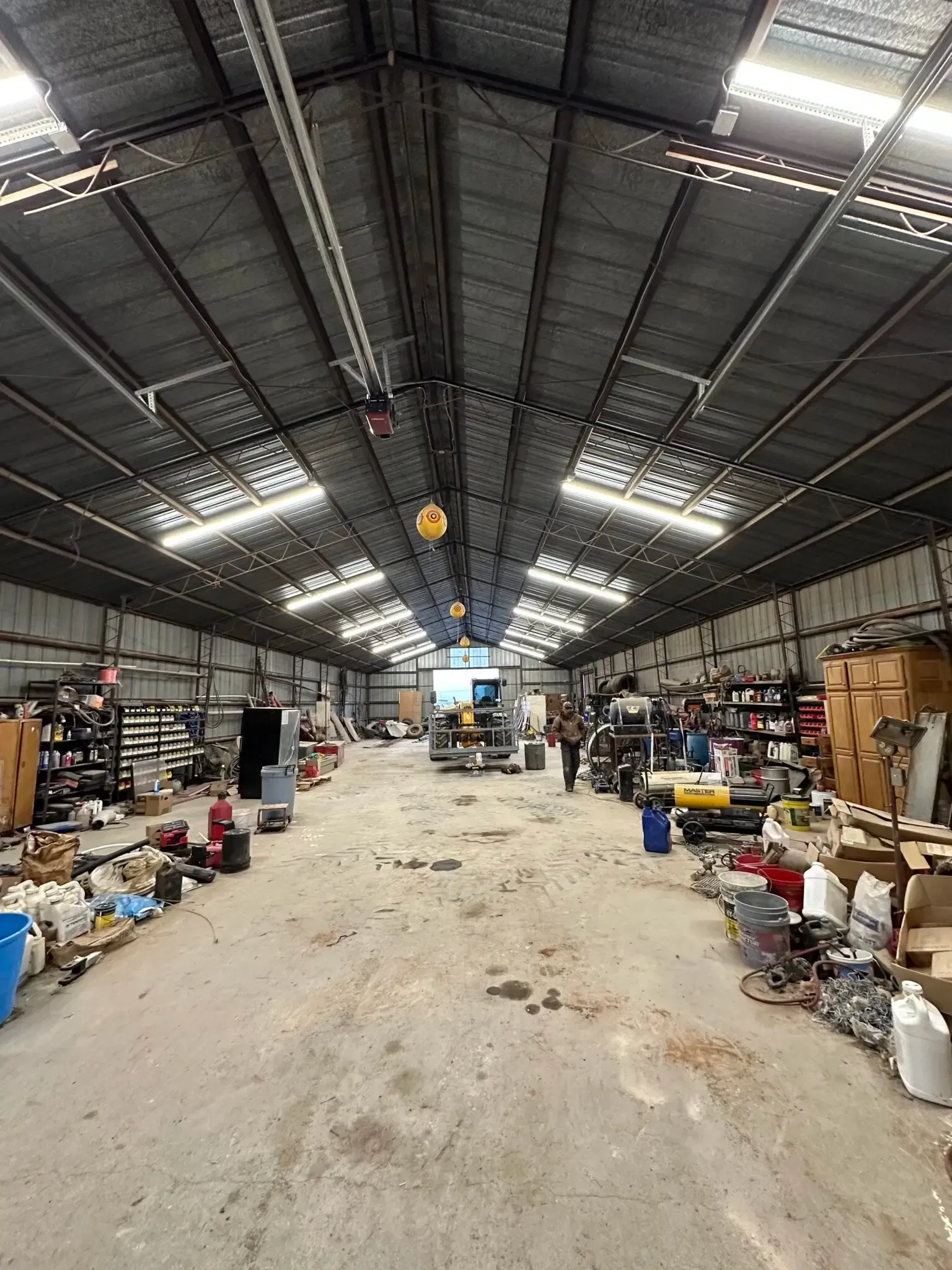 A wide-angle, eye-level view of a long, narrow industrial workshop with high ceilings, scattered tools, and equipment.