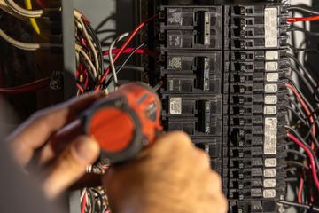 A person uses a red power drill to work on wires inside an open electrical circuit breaker panel.