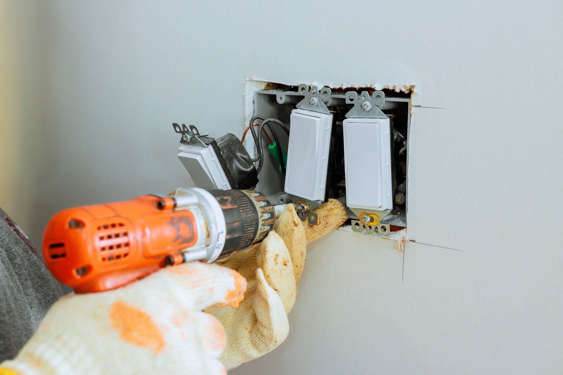 A person wearing work gloves uses an orange power drill to install a three-gang light switch panel in a wall.