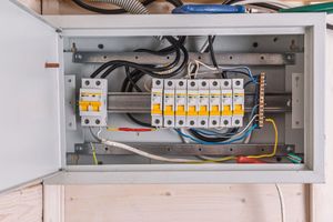 An open electrical service panel mounted on a wooden wall, showing circuit breakers and internal wiring.