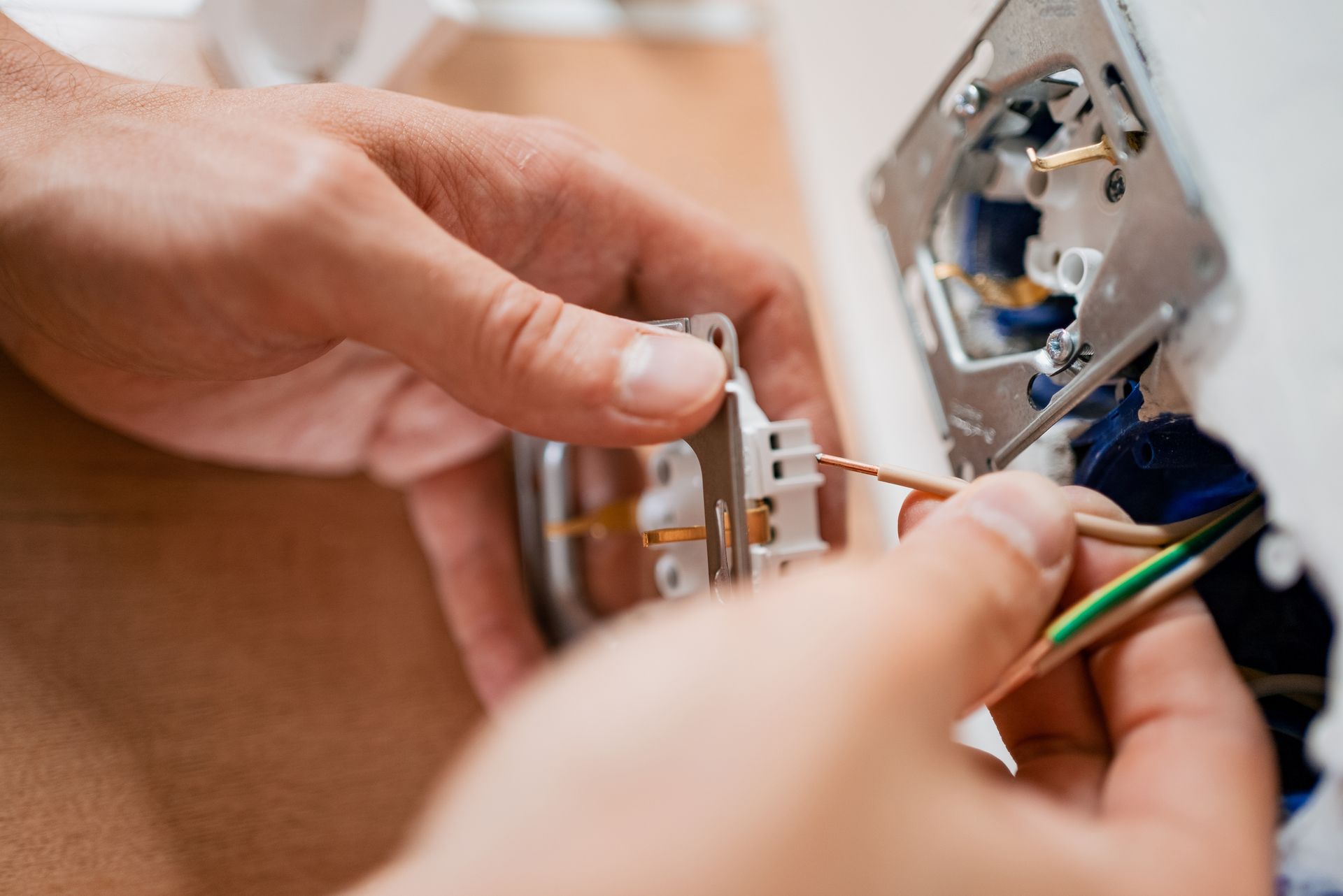 Hands wiring a wall electrical outlet, holding bare copper and green-and-yellow ground wires near a mounting bracket.