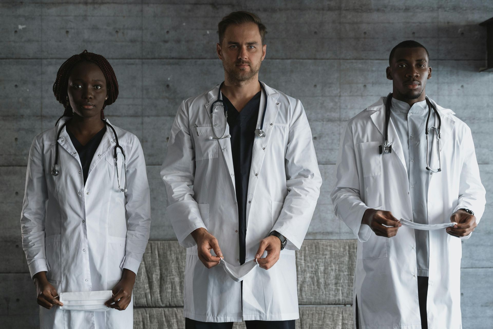 Three doctors in white coats, holding masks, stand in front of a concrete wall.