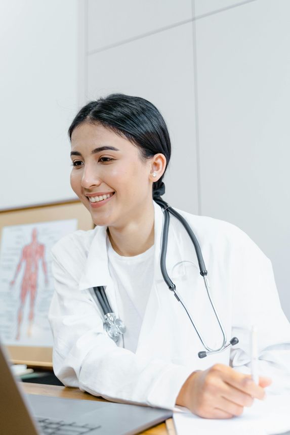 Smiling doctor with stethoscope at a desk, looking at a laptop and holding a glass.