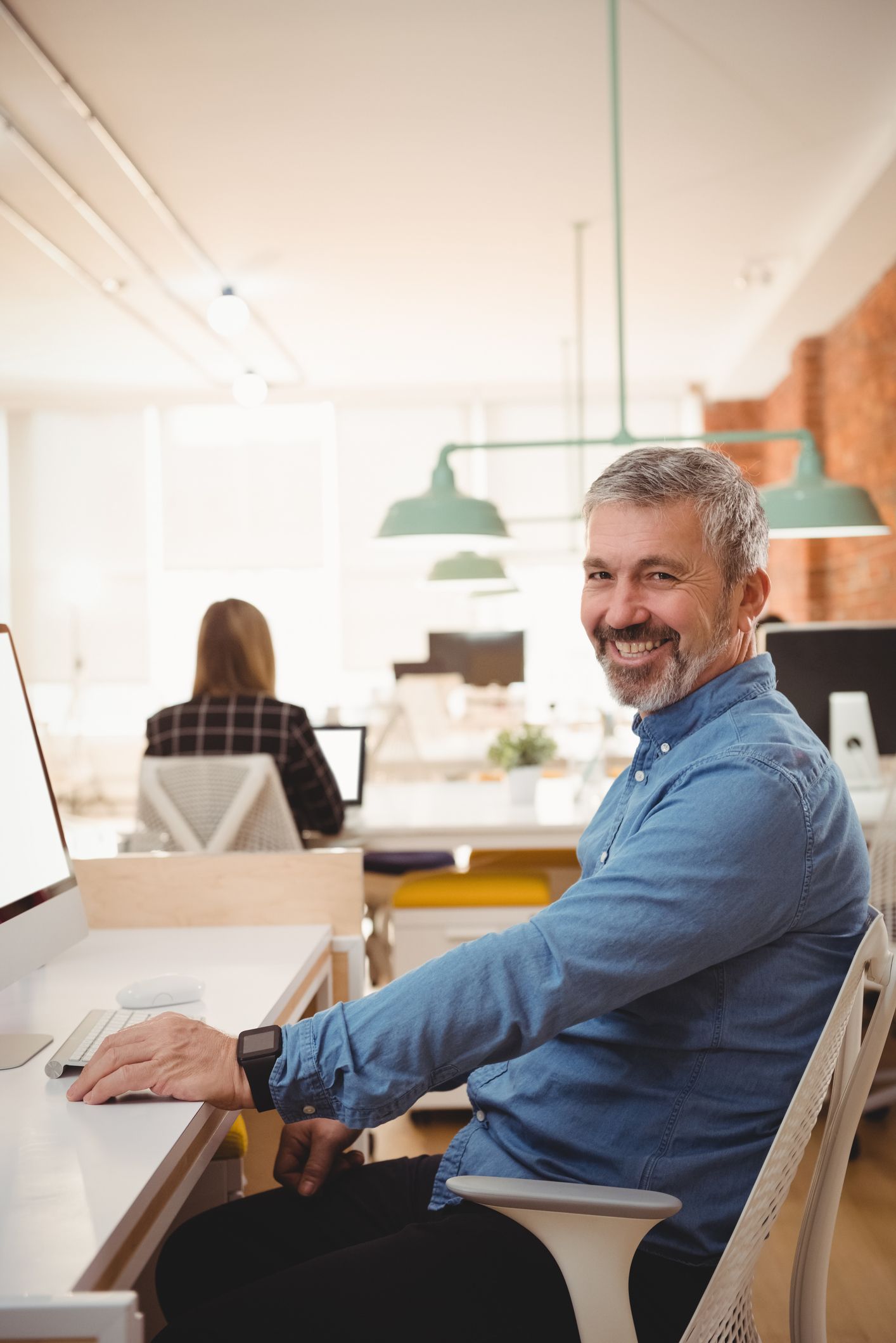 Man smiling, sitting at desk in office, looking at camera. Blue shirt, gray hair. Another person working in background.