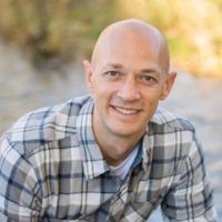 Bald man in a plaid shirt smiles outdoors. He's sitting, with a neutral background.